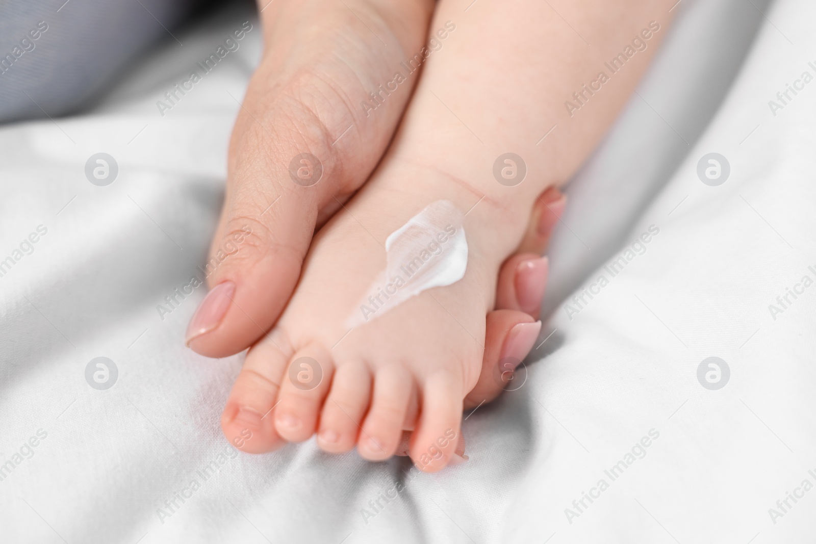 Photo of Woman applying body cream onto baby`s foot on bed, closeup