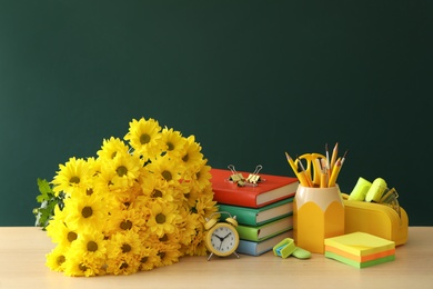 Set of stationery, alarm clock and flowers on wooden table near chalkboard, space for text. Teacher's Day