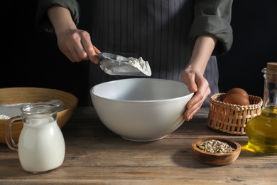 Making bread. Woman adding flour into bowl at wooden table on dark background, closeup