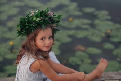 Cute little girl wearing wreath made of beautiful flowers near pond