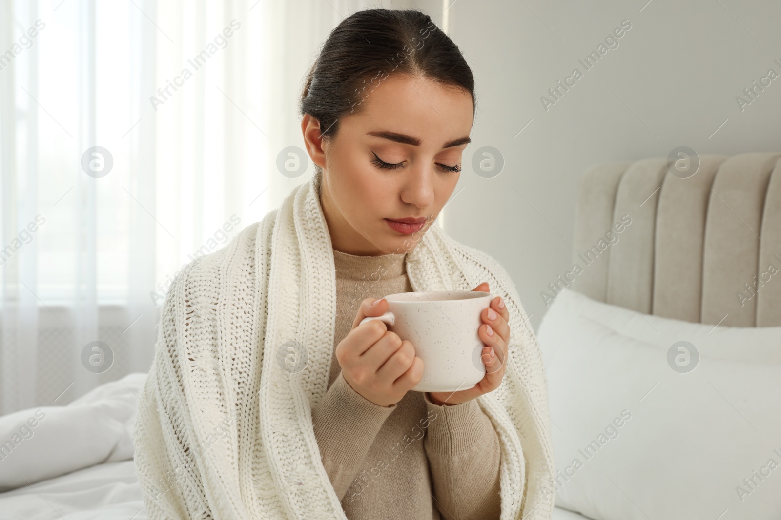 Photo of Beautiful young woman with cup of drink wrapped in knitted plaid at home