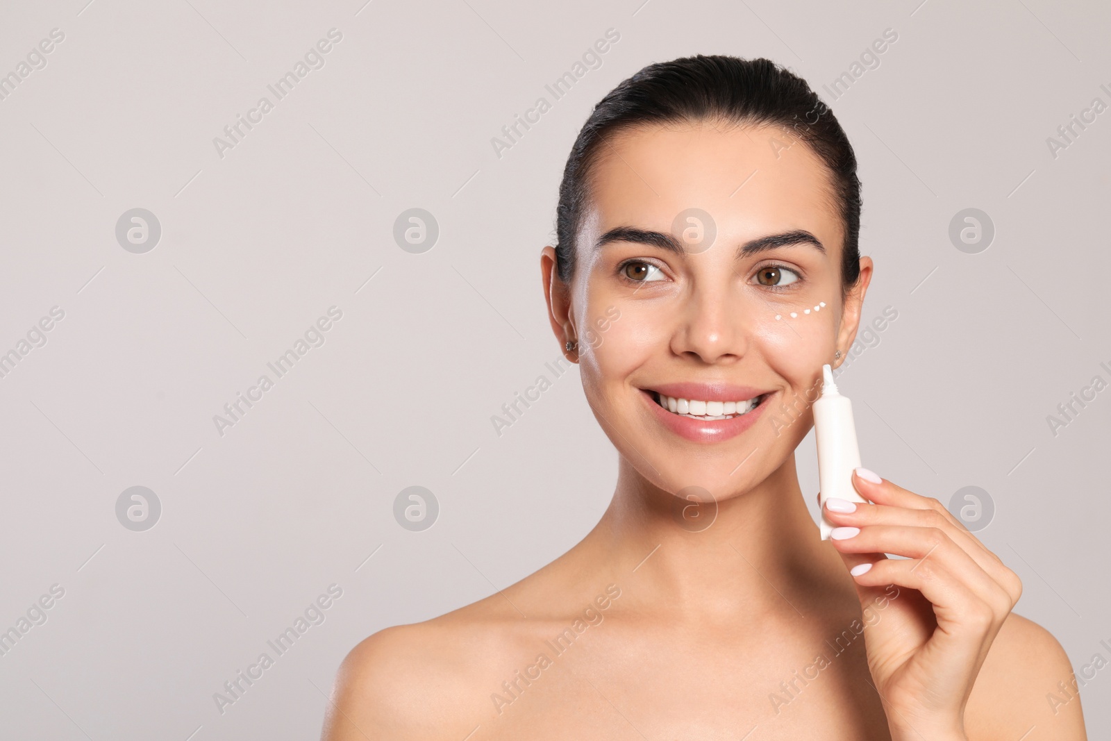 Photo of Beautiful young woman with cream on skin under eye against light grey background. Space for text