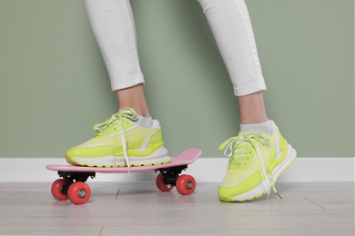 Photo of Woman in new stylish sneakers standing on skateboard near light green wall, closeup