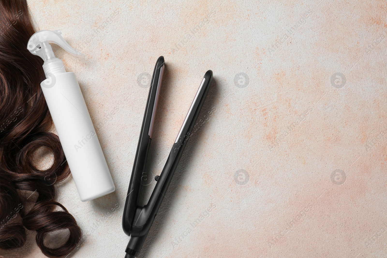 Photo of Spray bottle with thermal protection, lock of brown hair and modern straightener on beige textured table, flat lay. Space for text