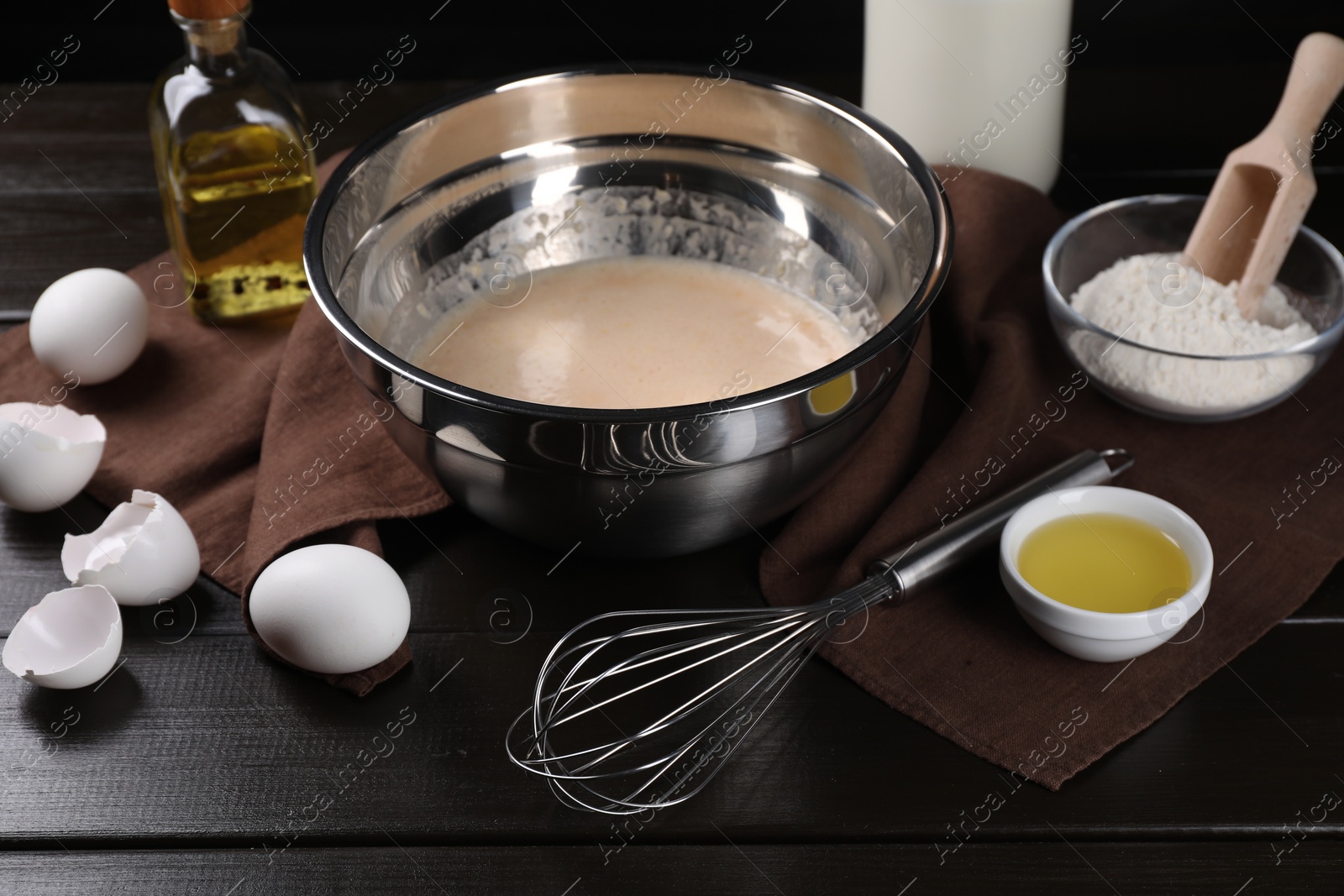 Photo of Dough in bowl, metal whisk and ingredients on dark wooden table, closeup