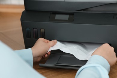 Photo of Woman taking crumpled paper out of printer at wooden table indoors, closeup