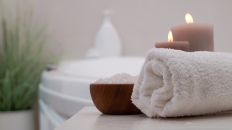 Photo of Rolled bath towel, sea salt and burning candles on table in bathroom, closeup. Space for text