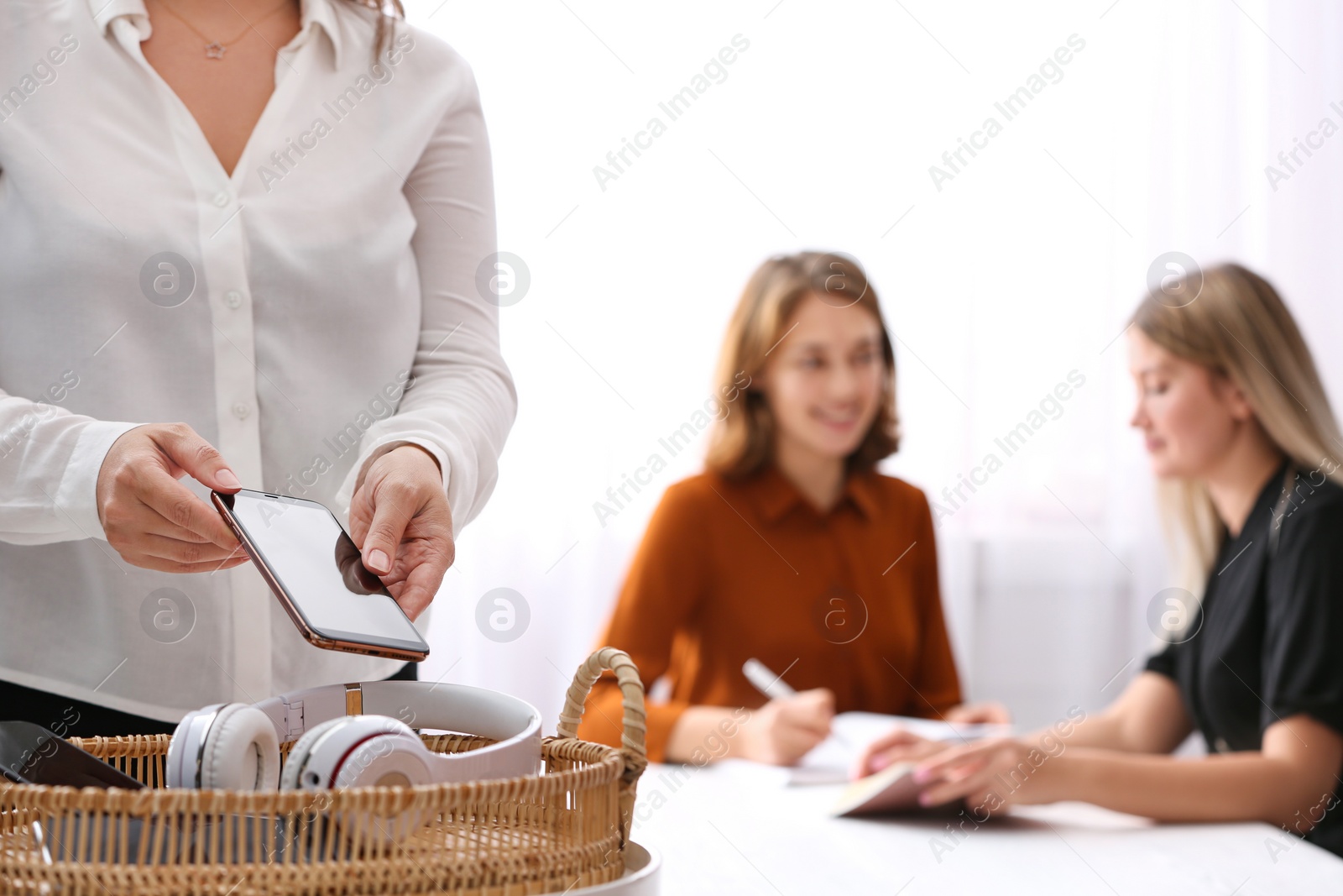 Photo of Woman putting smartphone into wicker basket with gadgets in office, closeup. Digital detox concept