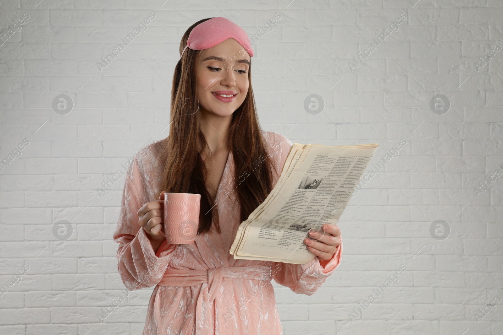 Photo of Beautiful young woman in bathrobe and eye sleeping mask reading newspaper near white brick wall