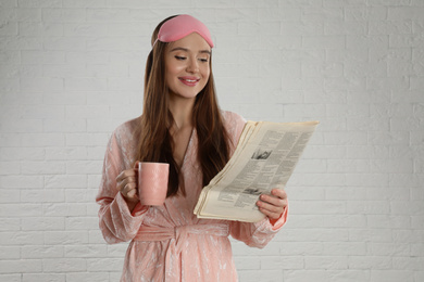 Photo of Beautiful young woman in bathrobe and eye sleeping mask reading newspaper near white brick wall