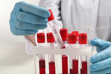 Scientist taking tube with blood sample and label STD Test from rack at white table, closeup