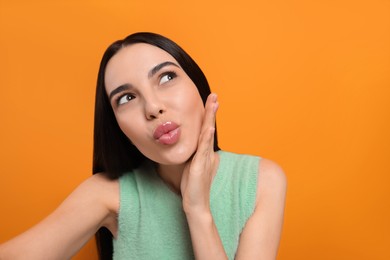 Beautiful young woman taking selfie while blowing kiss on orange background. Space for text