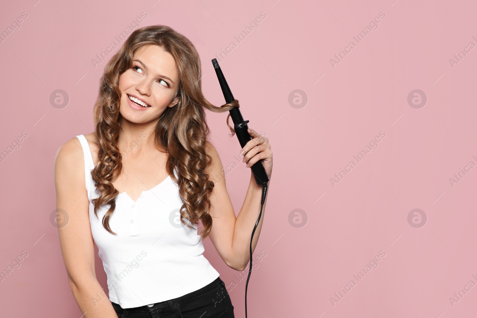 Photo of Portrait of young woman with shiny wavy hair using curling iron on color background. Space for text