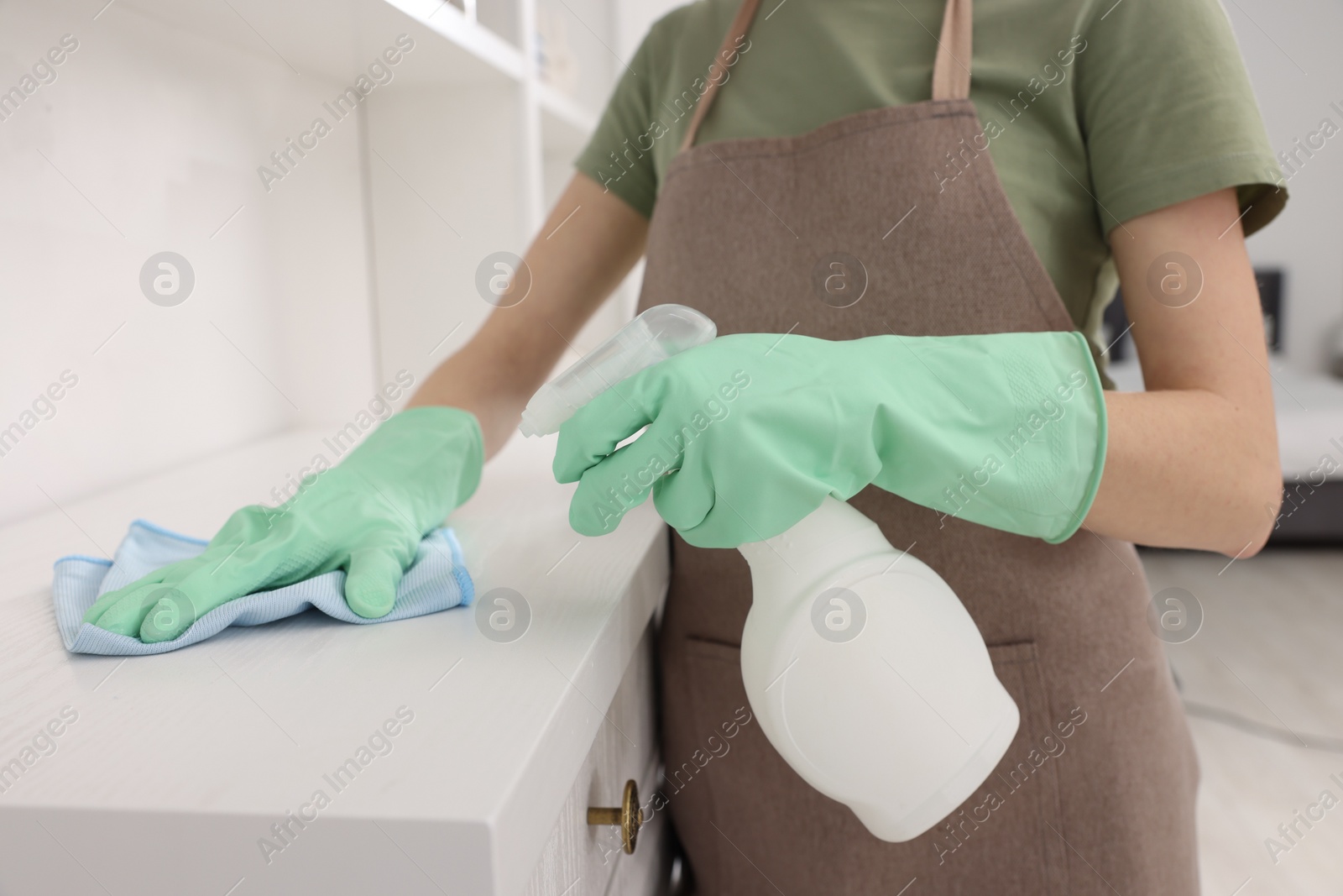 Photo of Woman with spray bottle and microfiber cloth cleaning white chest of drawers in room