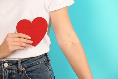 Woman holding heart near hand with adhesive plasters against color background, closeup. Blood donation