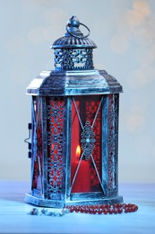 Photo of Arabic lantern and misbaha on table against blurred lights