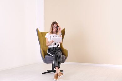 Young woman with tablet sitting in armchair at home. Space for text