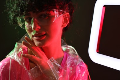 Photo of Beautiful young woman in transparent coat and sunglasses with square lamp on color background in neon lights
