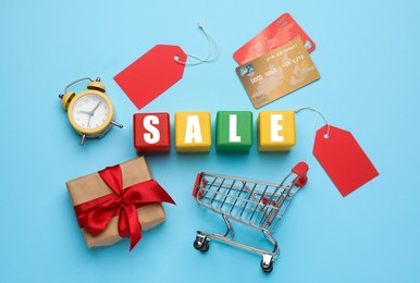 Photo of Wooden cubes with word Sale, shopping cart, gift, credit cards and alarm clock on light blue background, flat lay