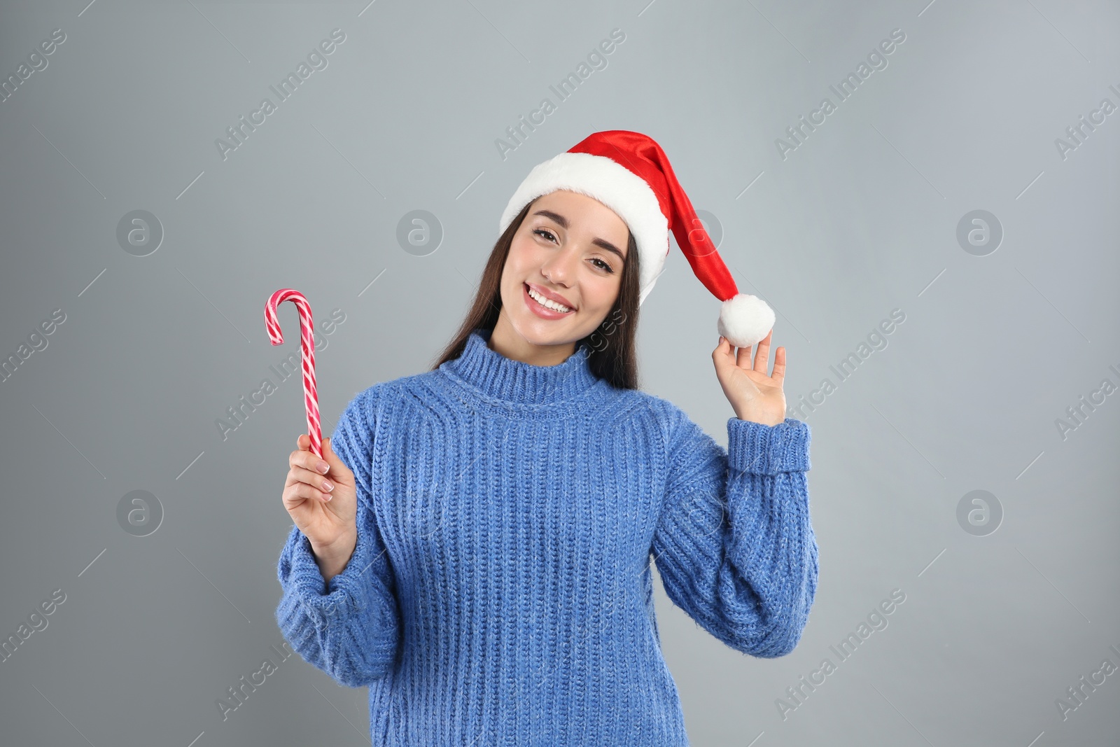 Photo of Young woman in blue sweater and Santa hat holding candy cane on grey background. Celebrating Christmas