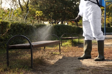 Photo of Person in hazmat suit spraying disinfectant onto bench outdoors, closeup. Surface treatment during coronavirus pandemic