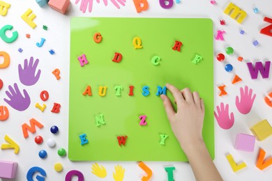 Photo of Woman making word Autism on magnetic board at white table, top view