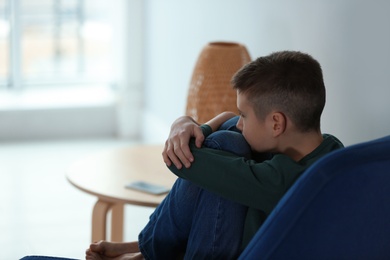 Upset boy sitting in armchair indoors. Space for text