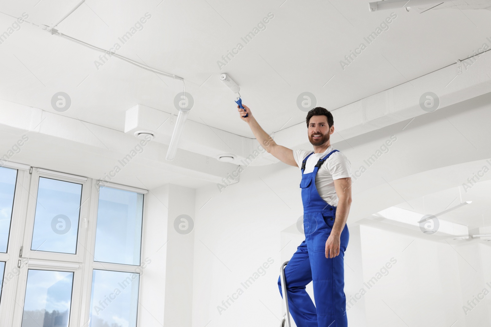 Photo of Handyman painting ceiling with roller on step ladder in room