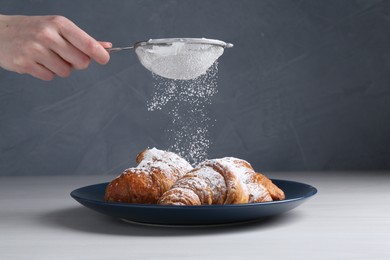Woman with sieve sprinkling powdered sugar onto croissants at white wooden table, closeup