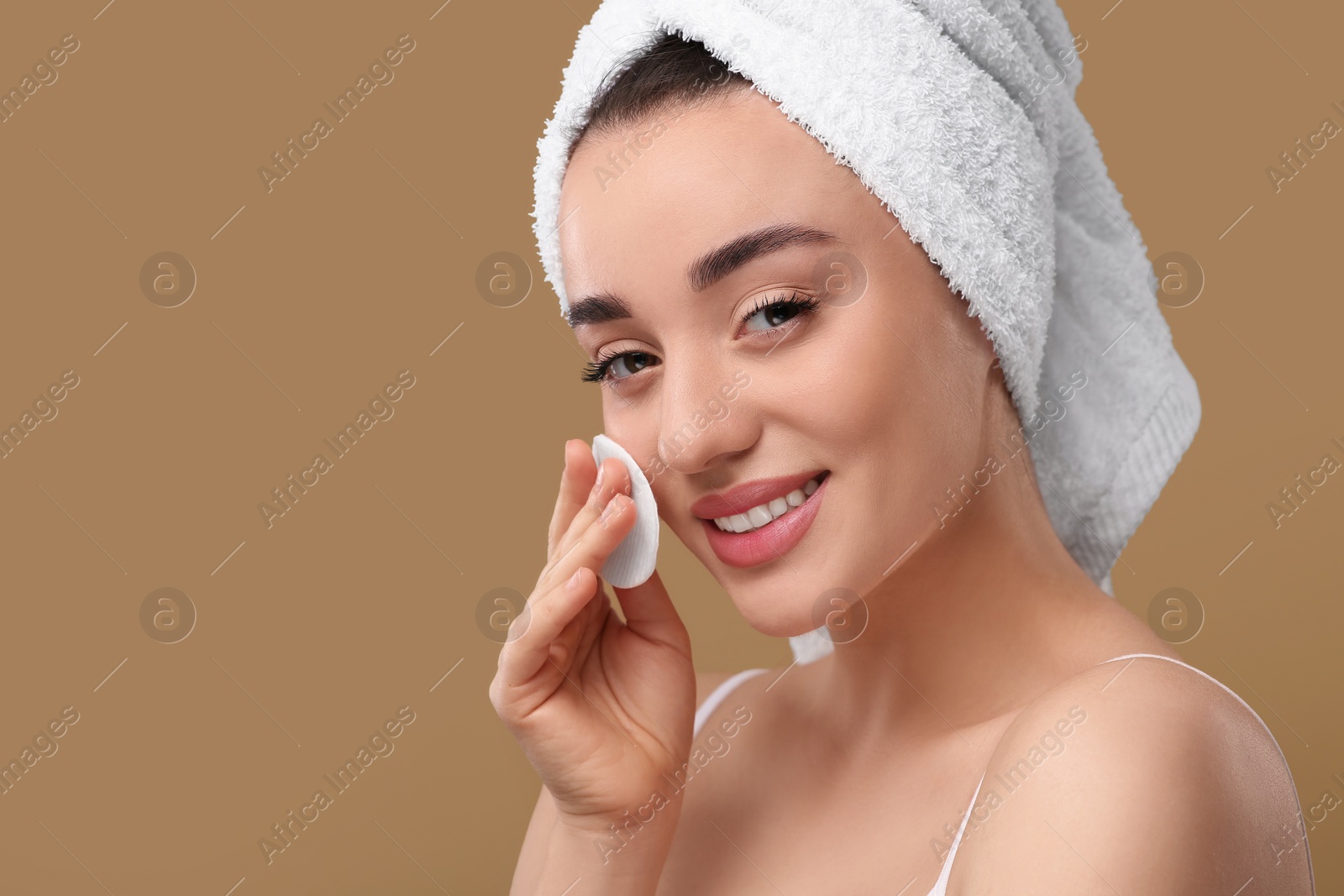 Photo of Beautiful woman in terry towel removing makeup with cotton pad on beige background, space for text