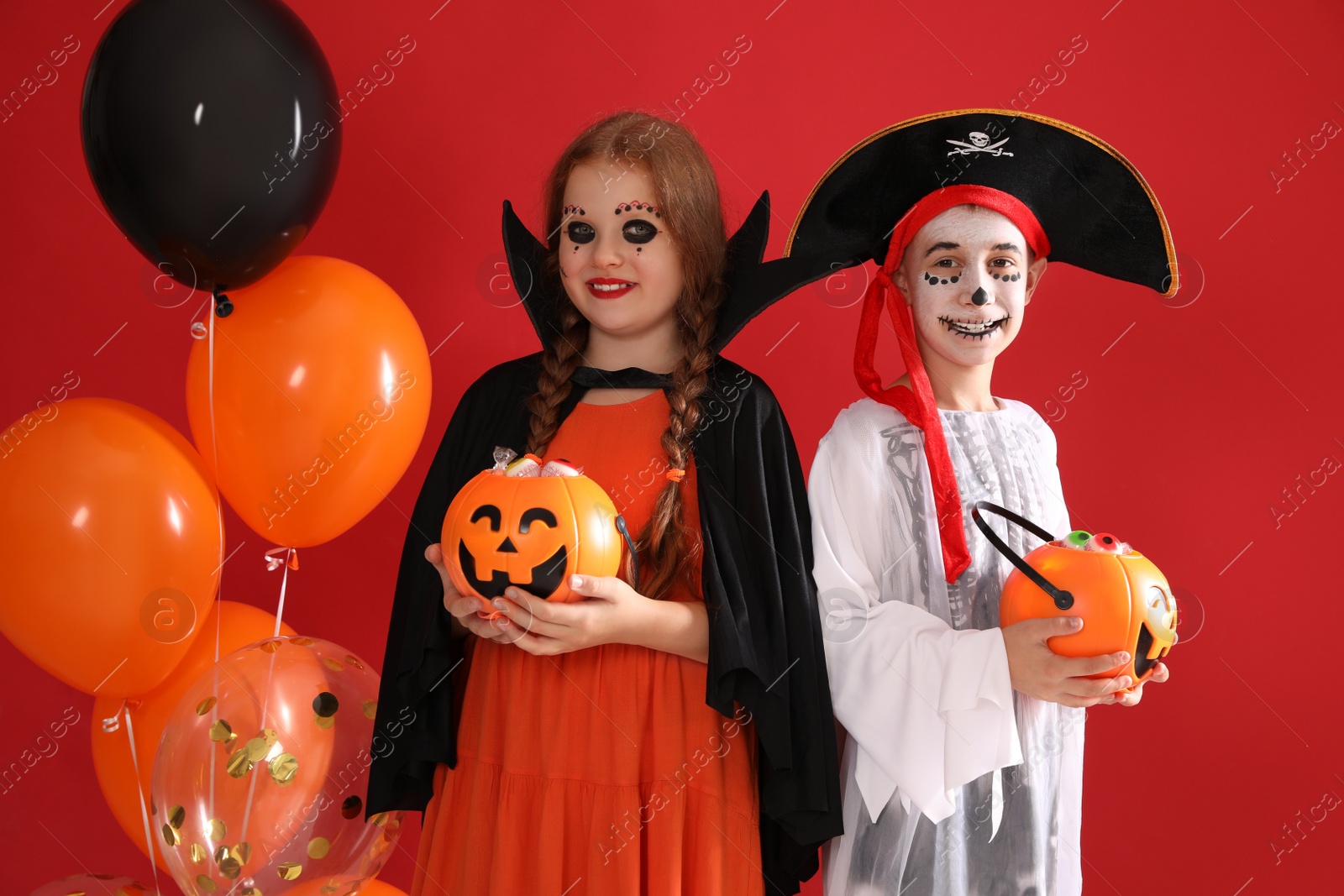 Photo of Cute little kids with pumpkin candy buckets and balloons wearing Halloween costumes on red background