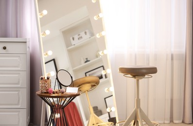 Makeup room. Stylish mirror with light bulbs, beauty products on table and chair indoors