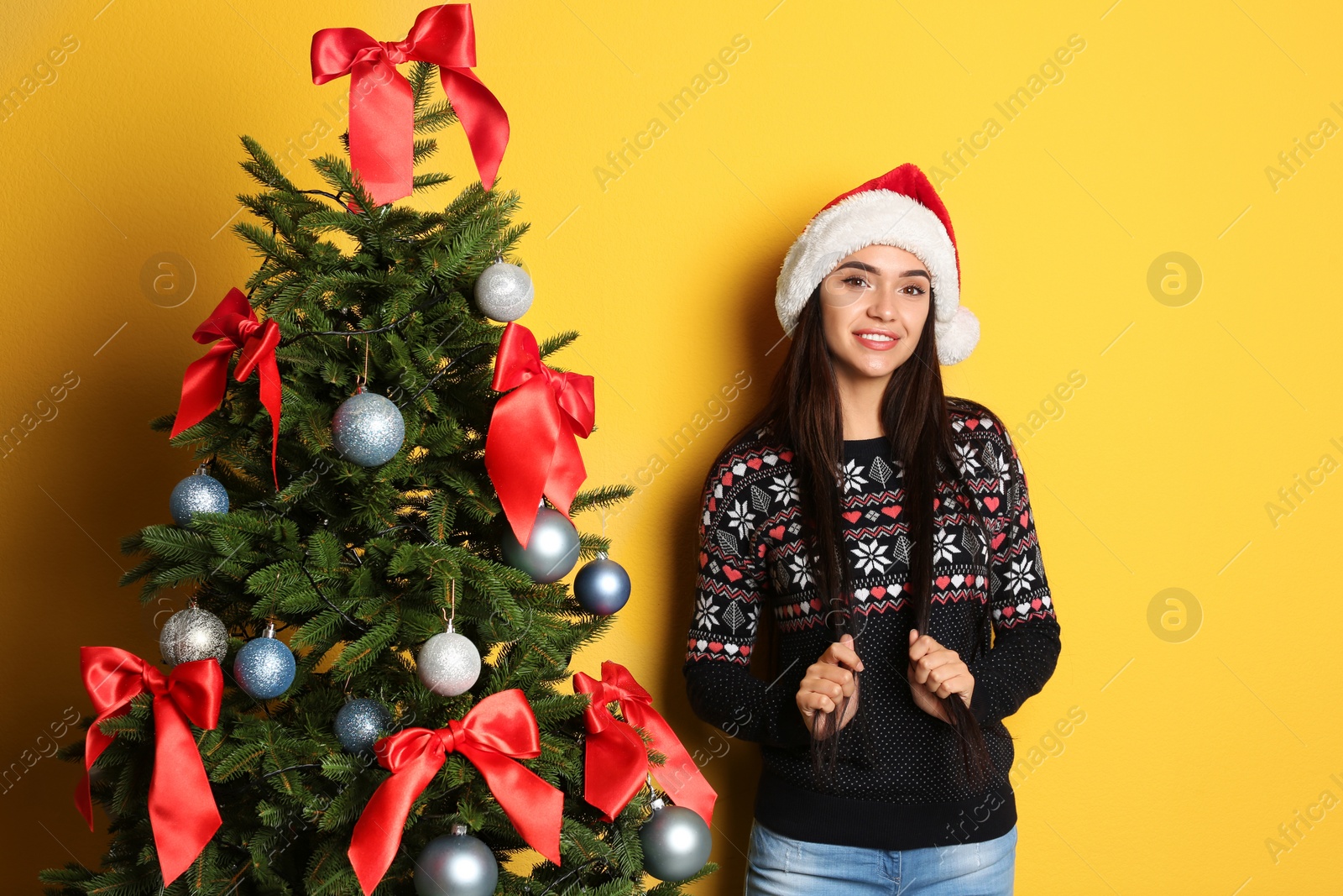 Photo of Beautiful young woman in Santa hat near Christmas tree on color background