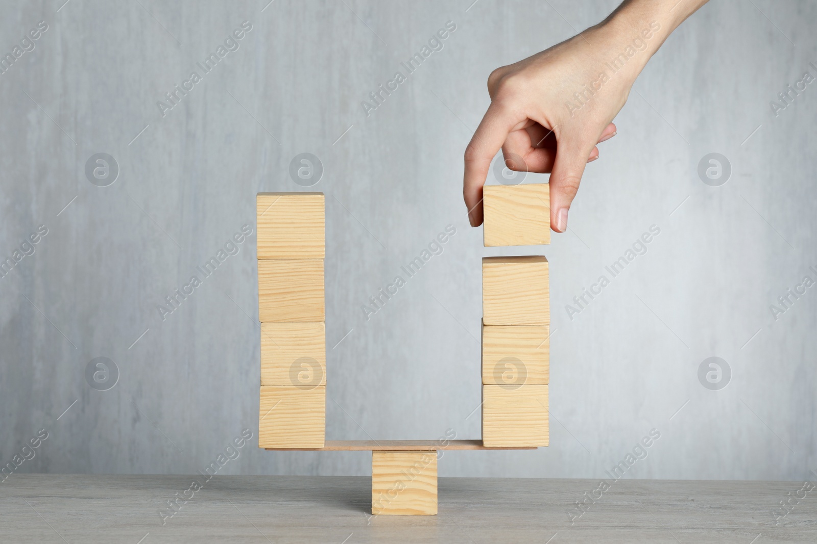 Photo of Woman balancing miniature seesaw with stacks of wooden cubes on table against light background, closeup. Harmony concept