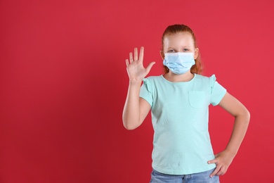 Photo of Little girl in protective mask showing hello gesture on red background, space for text. Keeping social distance during coronavirus pandemic