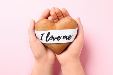 Woman holding wooden heart and paper with phrase I Love Me on pink background, top view