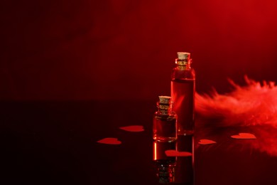 Bottles of love potion, paper hearts and feather on mirror surface against dark background, space for text. Red tone effect