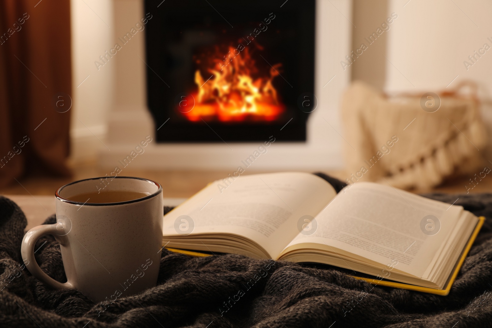 Photo of Cup of hot tea and book on knitted plaid near fireplace at home. Cozy atmosphere