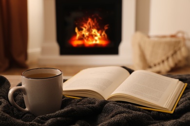 Cup of hot tea and book on knitted plaid near fireplace at home. Cozy atmosphere