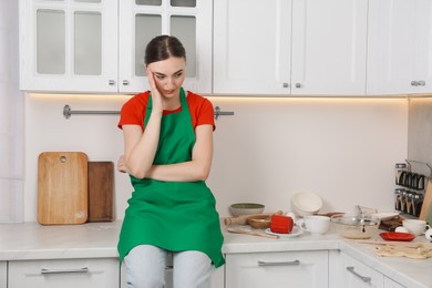 Upset woman sitting on countertop with dirty dishware and utensils. Mess in kitchen