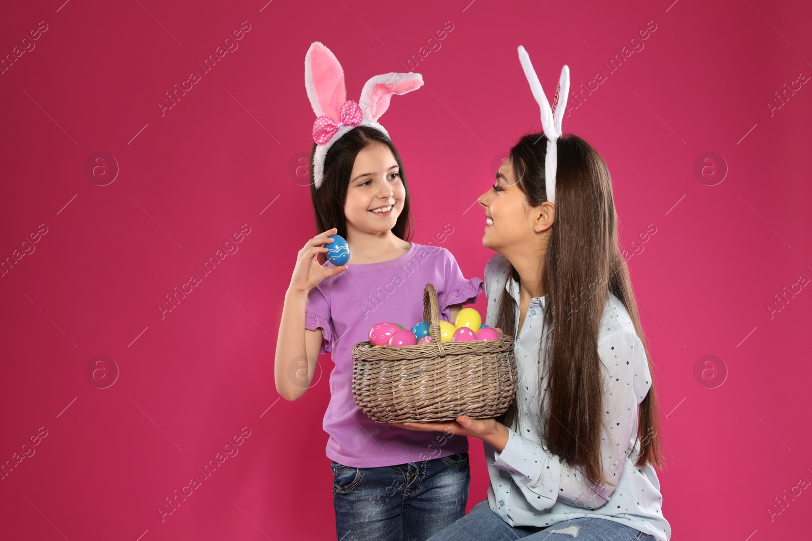 Photo of Mother and daughter in bunny ears headbands with basket of Easter eggs on color background