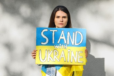 Photo of Sad woman holding poster in colors of national flag and words Stand with Ukraine near light wall