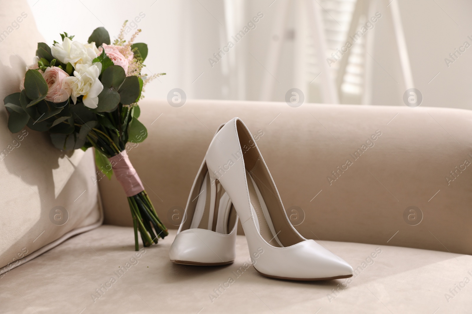 Photo of Pair of white wedding high heel shoes and beautiful bouquet on sofa indoors