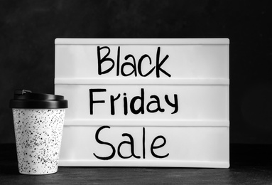 Photo of Reusable coffee cup and lightbox with words Black Friday Sale on table against dark background