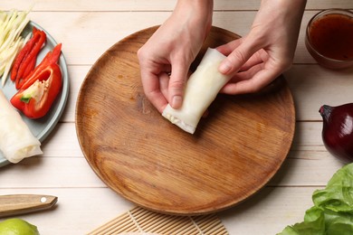 Photo of Making delicious spring rolls. Woman wrapping fresh vegetables into rice paper at wooden table, closeup