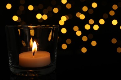 Glass holder with burning tea candle against blurred lights in darkness, closeup. Space for text