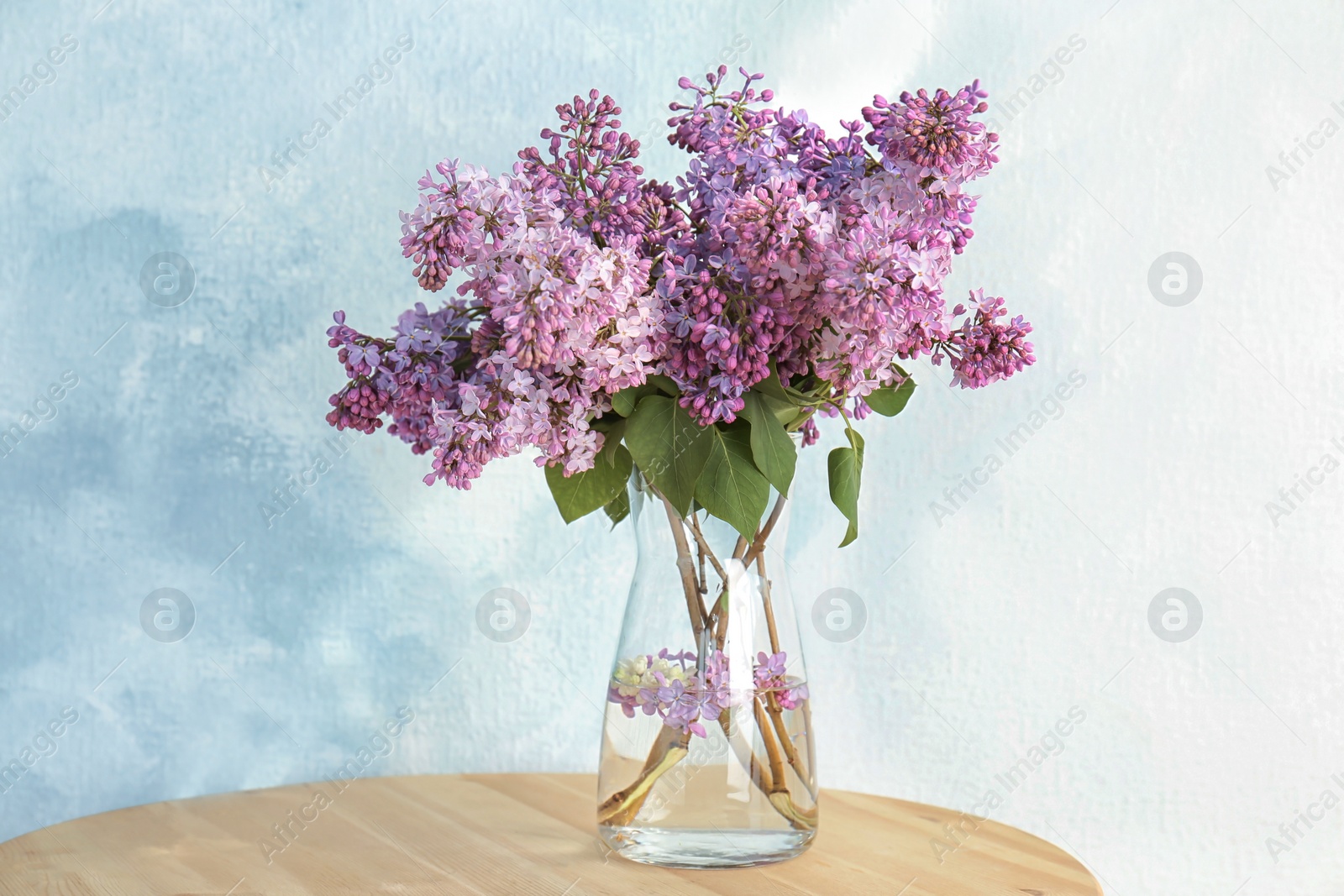 Photo of Vase with beautiful blossoming lilac on table against light background. Spring flowers