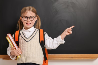 Photo of Happy schoolgirl in glasses with backpack and books pointing at something near blackboard, space for text