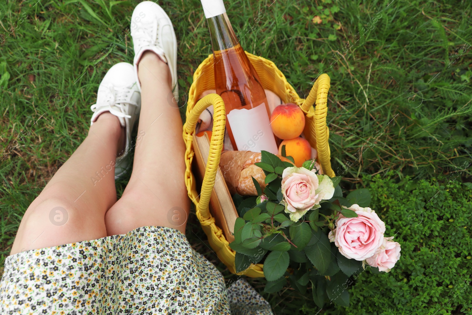 Photo of Woman sitting near yellow wicker bag with roses, wine, peaches and baguette on green grass outdoors, above view