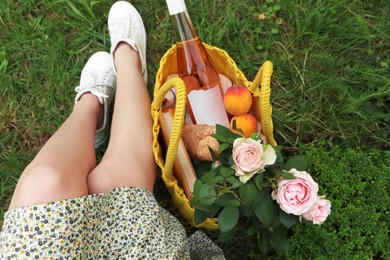 Woman sitting near yellow wicker bag with roses, wine, peaches and baguette on green grass outdoors, above view